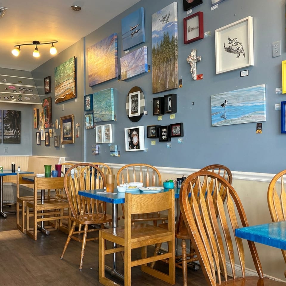 Bright restaurant dining room with blue tables, wooden chairs, and walls covered in colorful artwork.