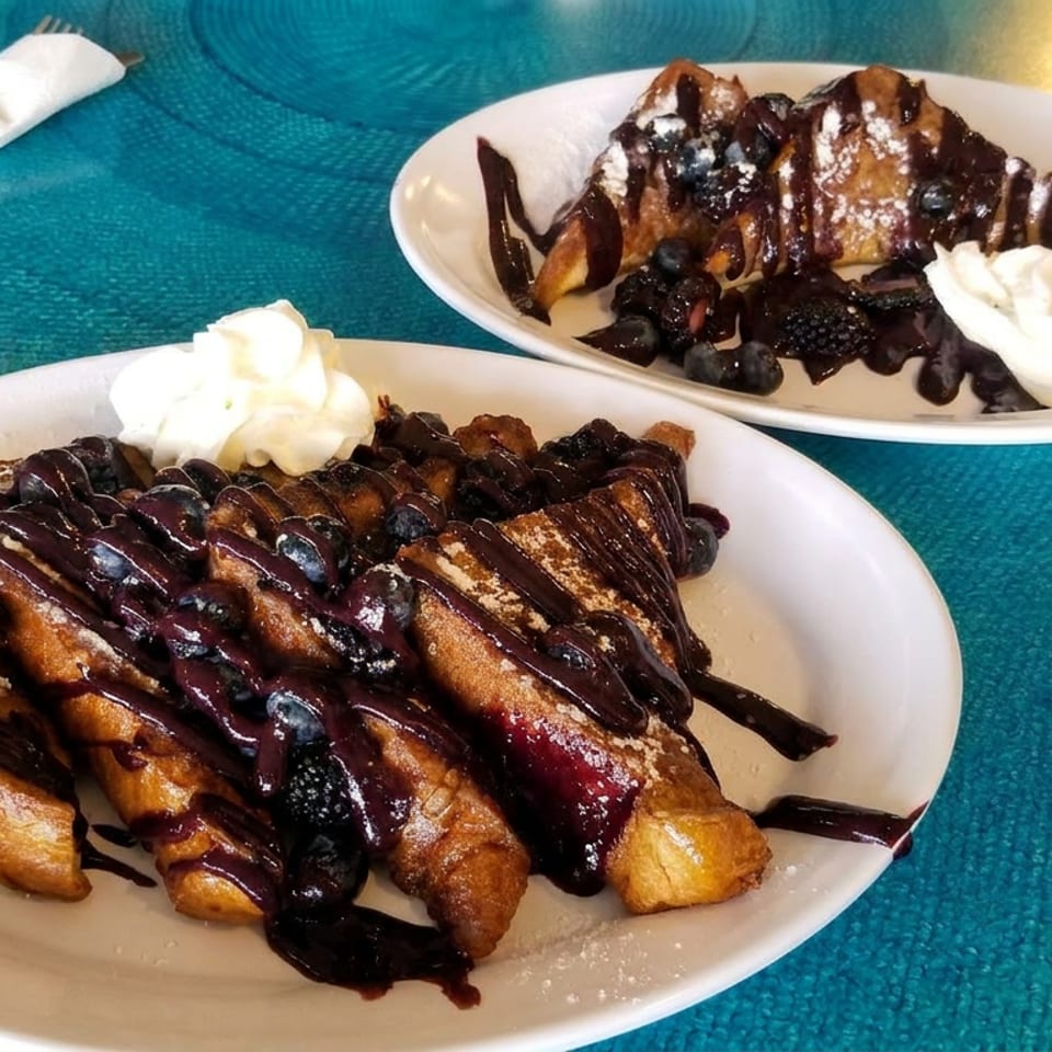 Two plates of golden dessert pastries drizzled with chocolate, topped with blueberries, powdered sugar, and whipped cream.