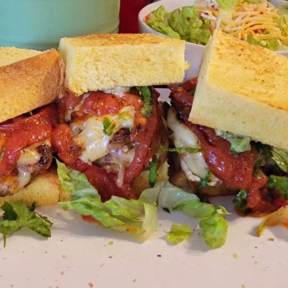 Toasted sandwich halves filled with meat, melted cheese, tomato, lettuce, and red sauce, served with a side salad.