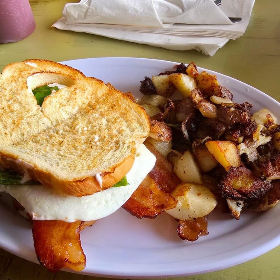 Toasted breakfast sandwich with fried egg, bacon, lettuce, and tomato served alongside crispy potatoes and onions.